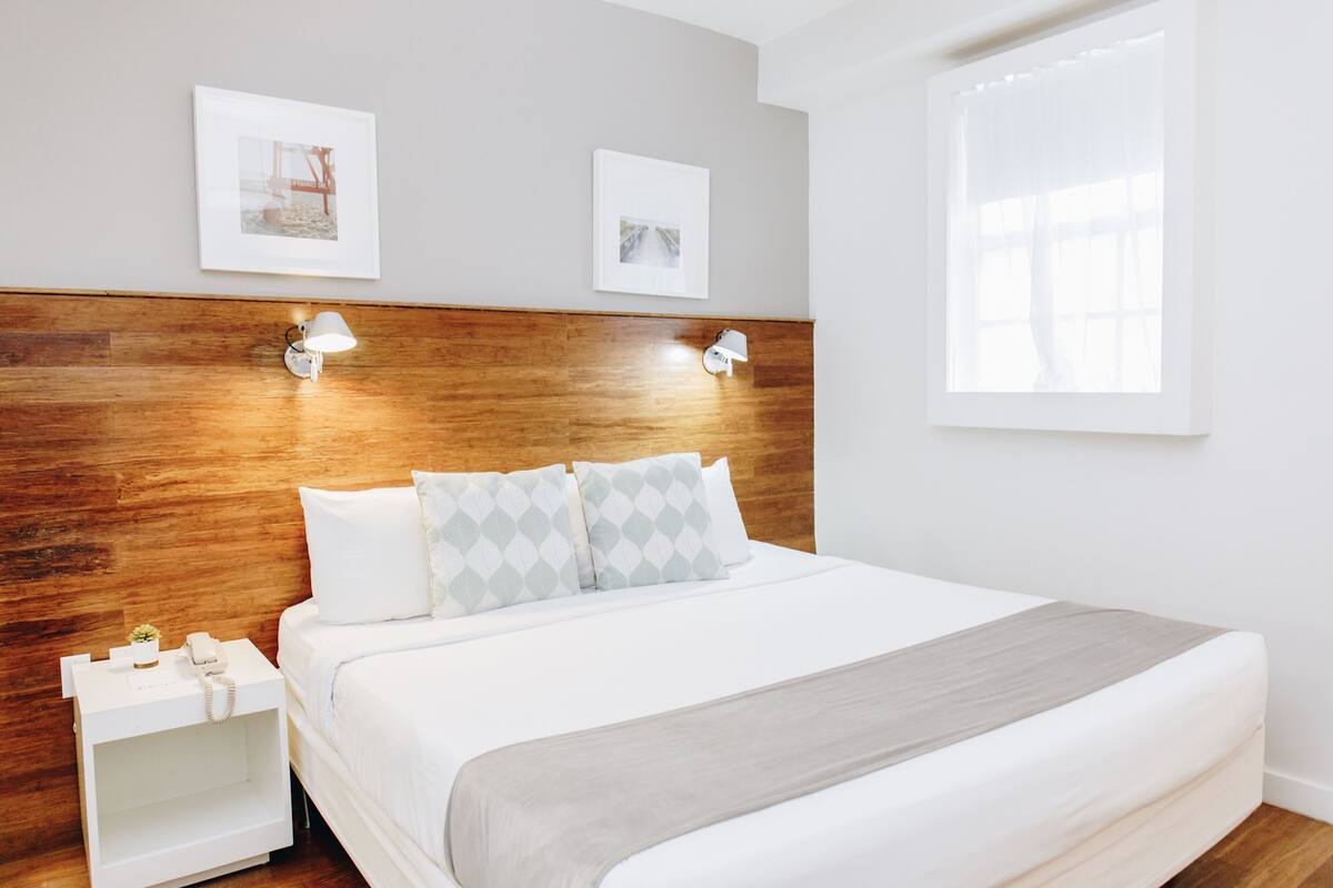 A neatly arranged hotel room with a wooden headboard, two bedside lamps, a bed with a grey blanket, and two framed pictures on the wall.