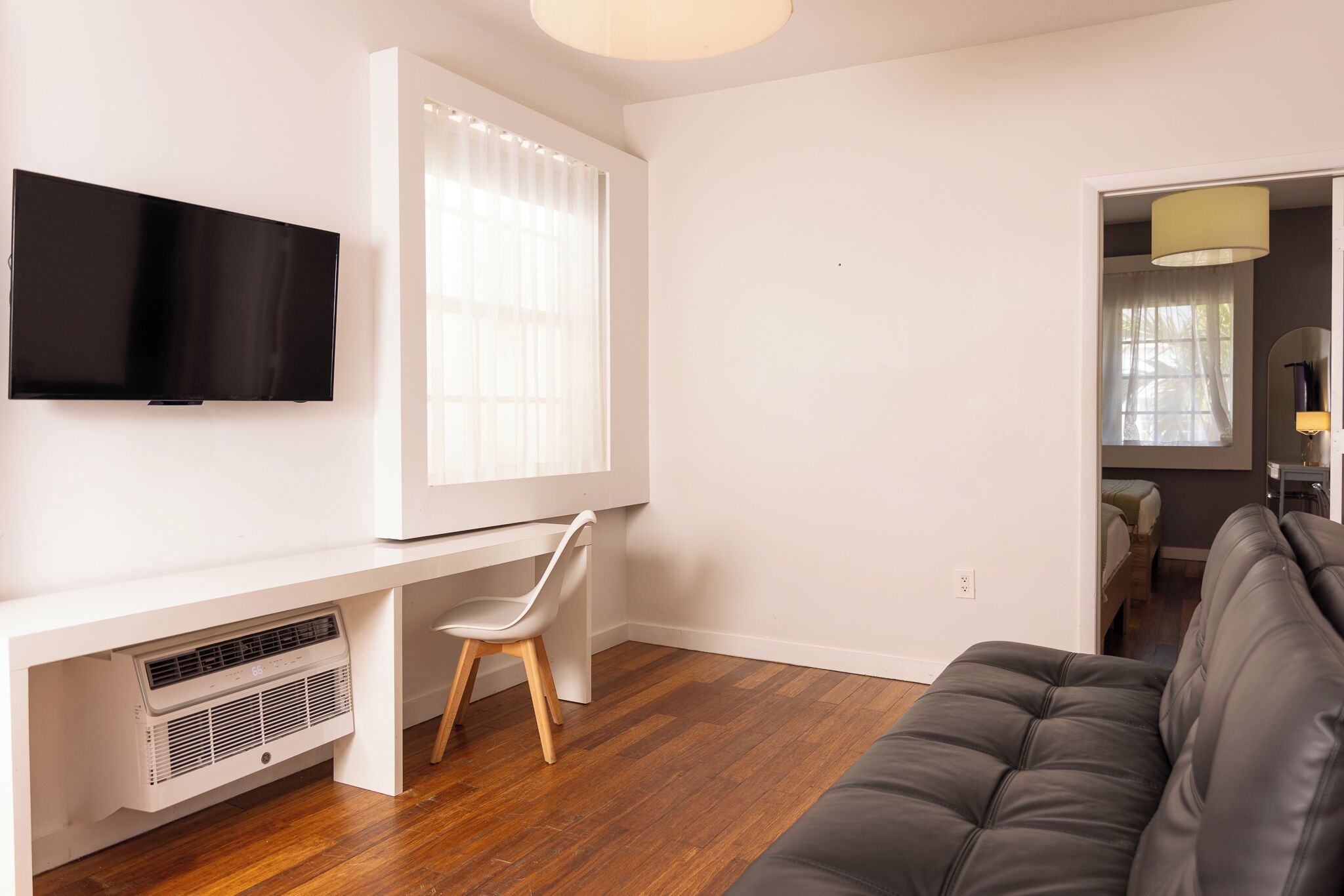 Un salon moderne avec un téléviseur à écran plat, un bureau blanc avec une chaise, un canapé gris et une fenêtre avec des stores.