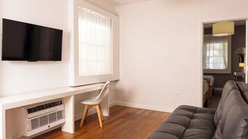 Un salon moderne avec un téléviseur à écran plat, un bureau blanc avec une chaise, un canapé gris et une fenêtre avec des stores.