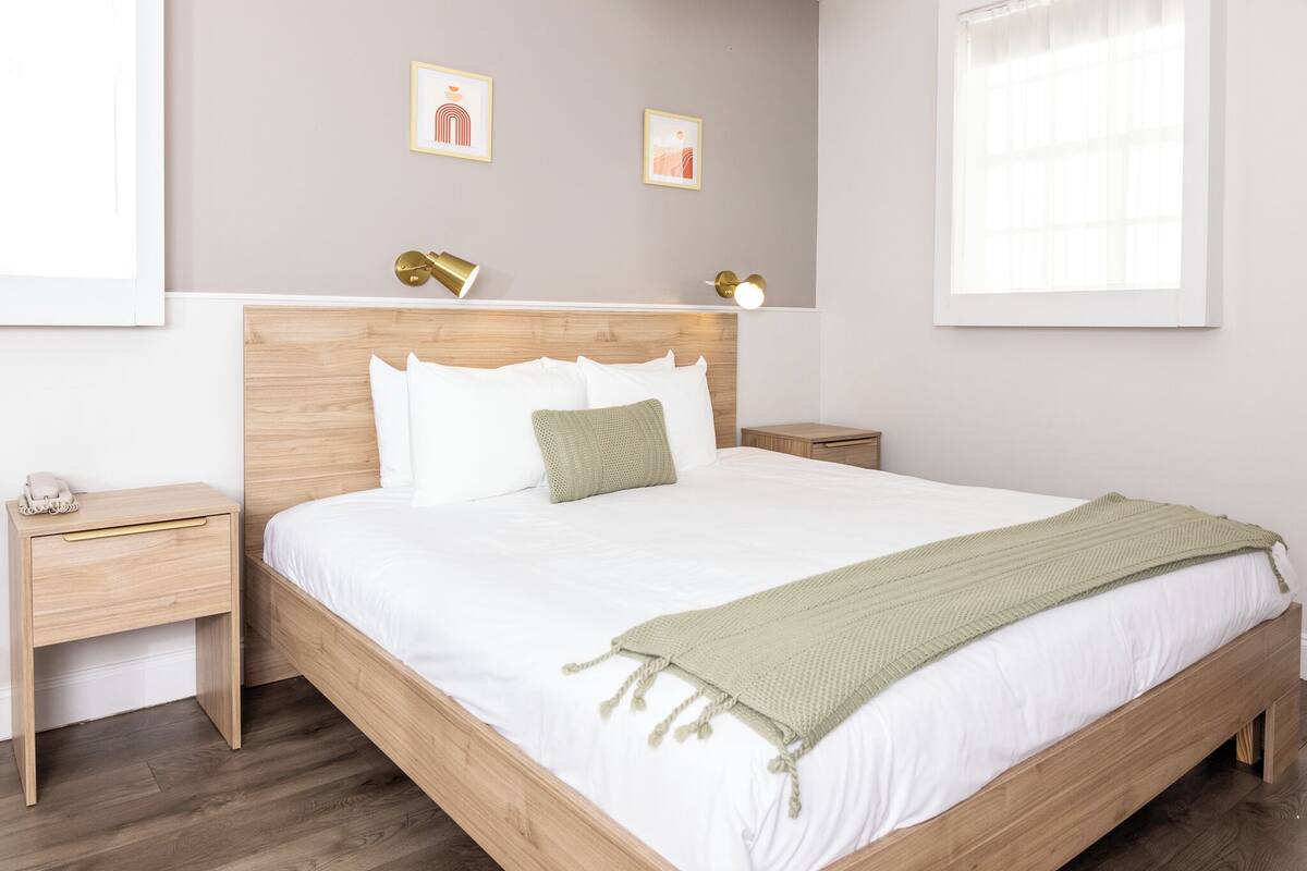 A neatly arranged bedroom with a wooden bed, white bedding, and a green throw blanket. There are two bedside tables with lamps, and artwork is displayed on the wall.