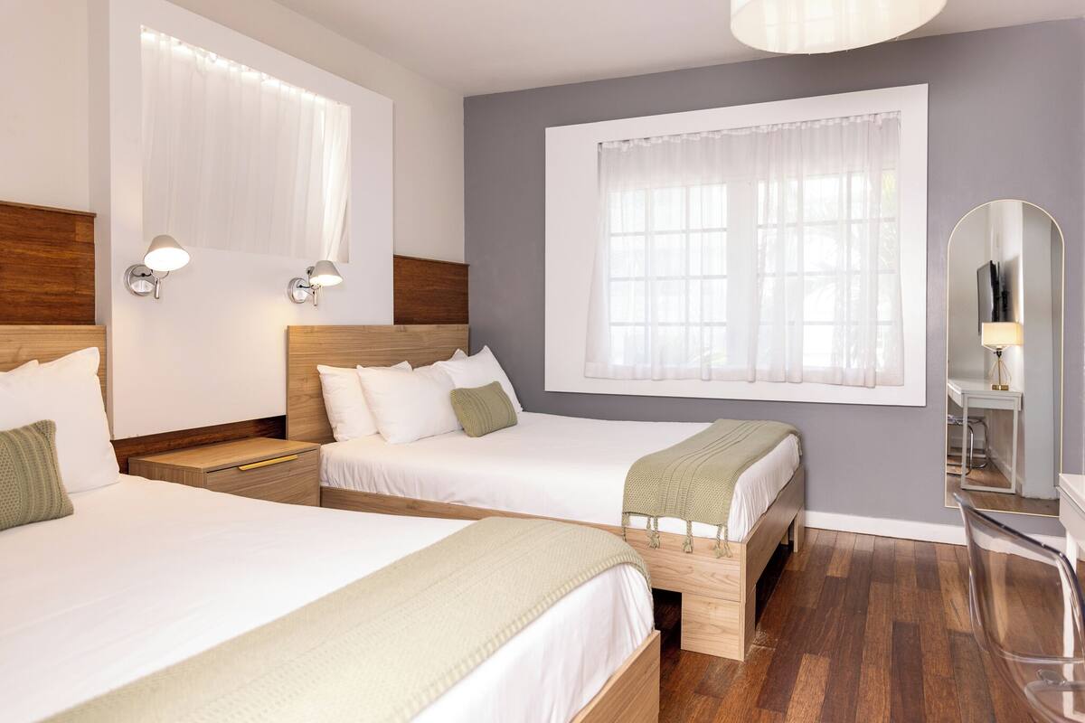 A hotel room with two single beds, wooden headboards, white bedding, a large window with sheer curtains, and a mirror on the wall.