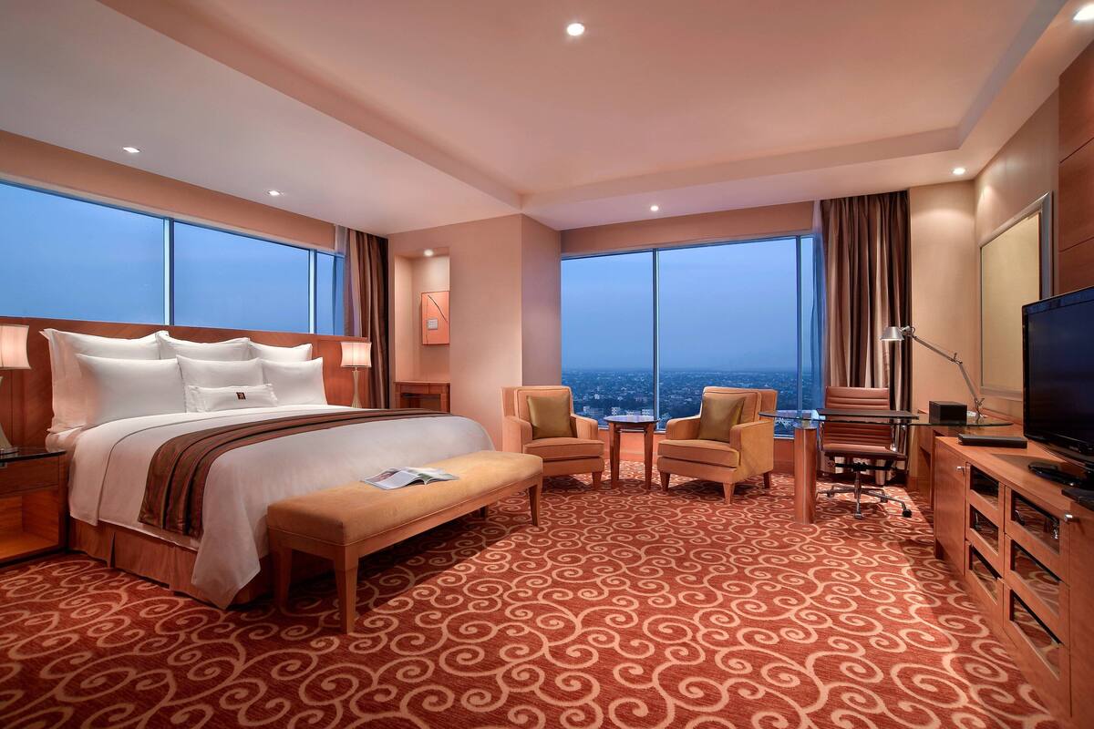 A hotel room with a large bed, a desk, two chairs, a television, and large windows offering a city view.