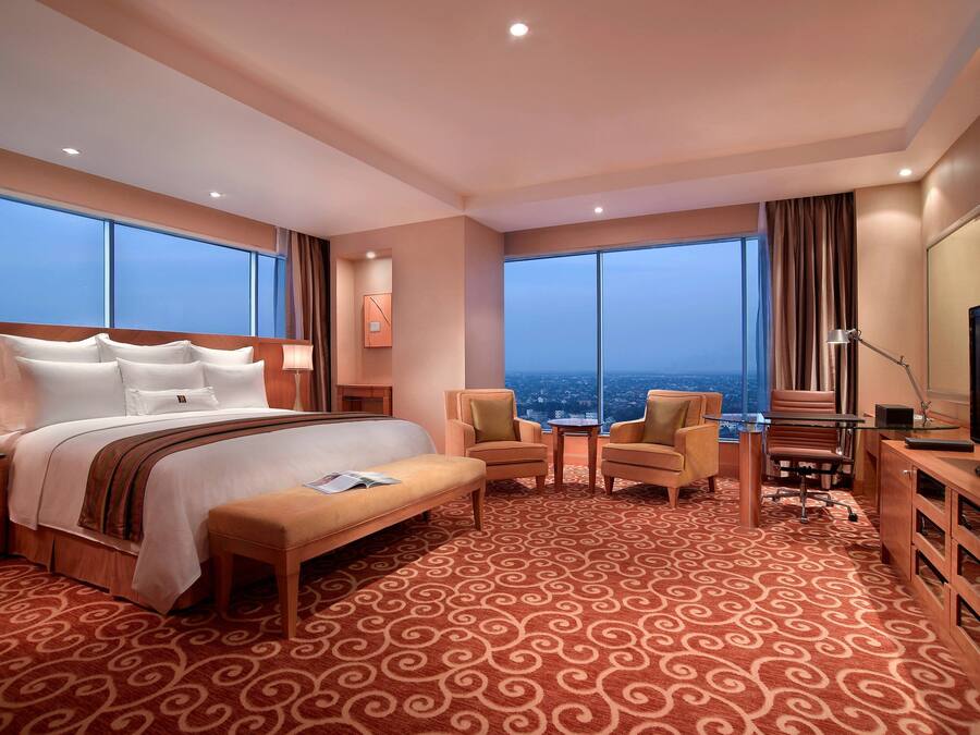 A hotel room with a large bed, a desk, two chairs, a television, and large windows offering a city view.