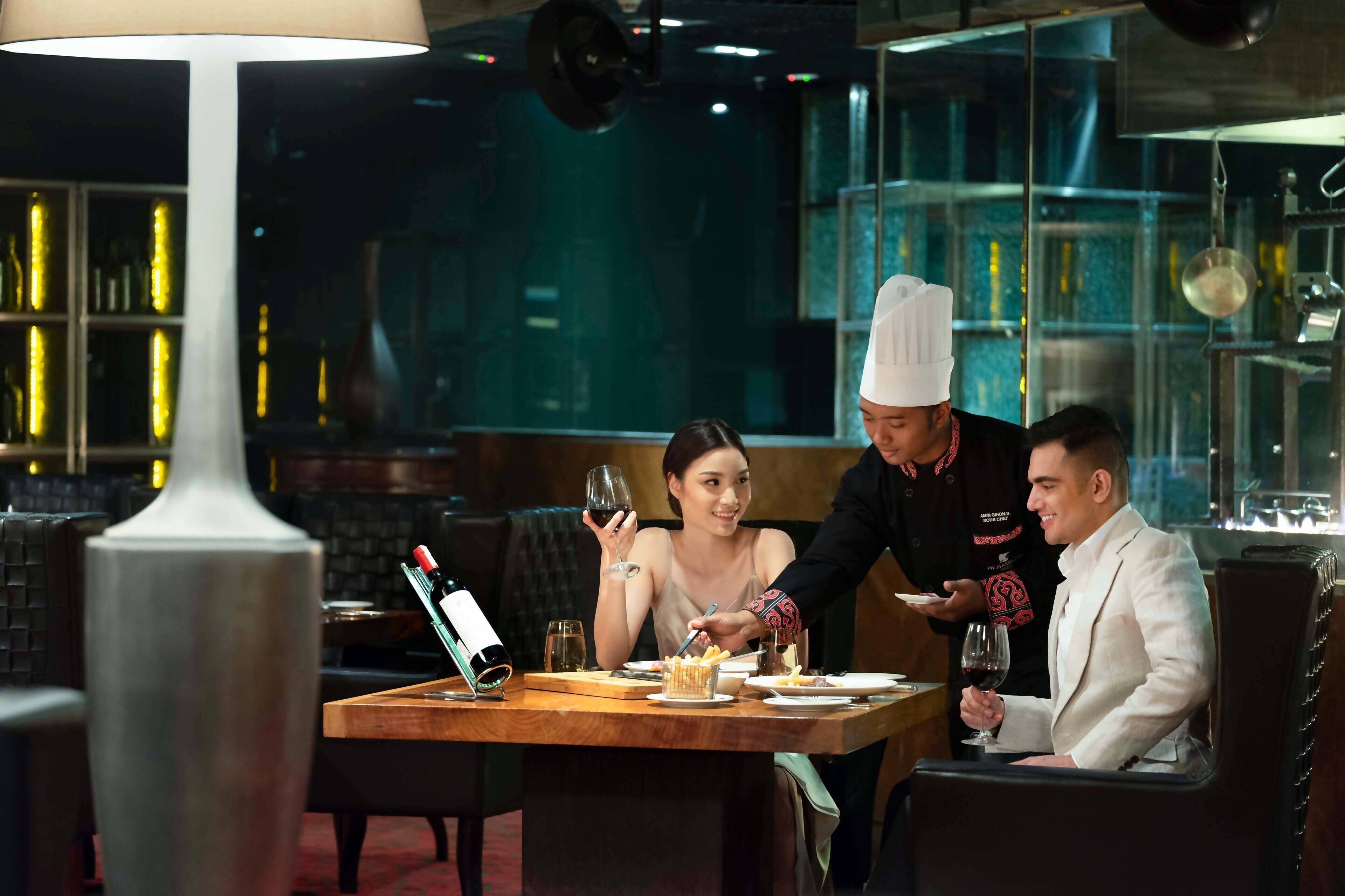 A restaurant setting with a table, chairs, a bottle of wine, and a person serving food.