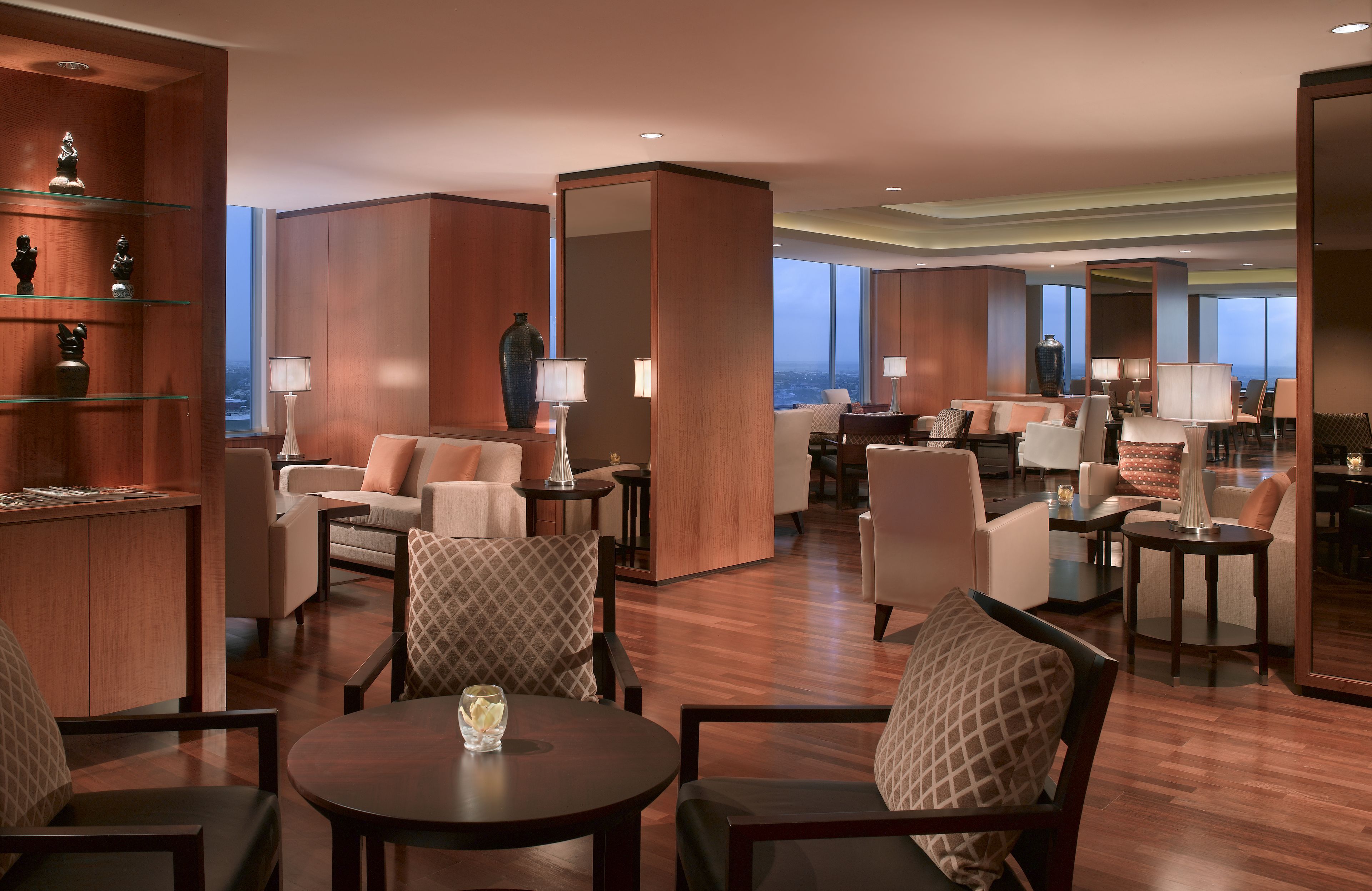 Un amplio vestíbulo de hotel con techo alto, paneles de madera y vistas a la ciudad. Varias áreas de estar con mesas y sillas, y un mostrador de recepción con vitrina.