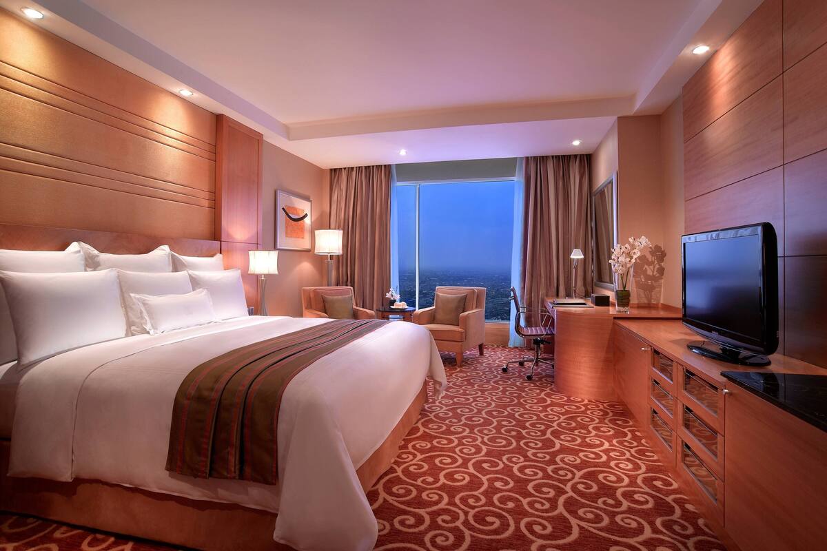 A hotel room with a large bed, a television, a desk with a chair, and a view of the ocean through the window.
