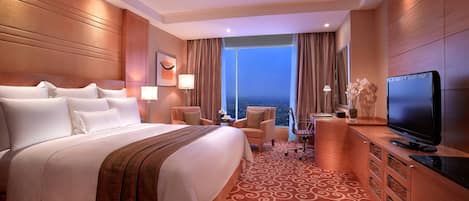 A hotel room with a large bed, a television, a desk with a chair, and a view of the ocean through the window.