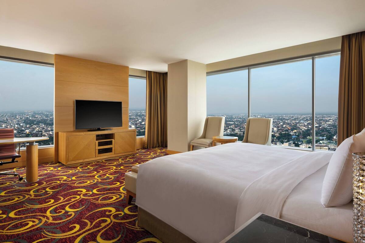 A hotel room with a large bed, a TV mounted on the wall, a desk, and a chair. The room has large windows offering a city view.