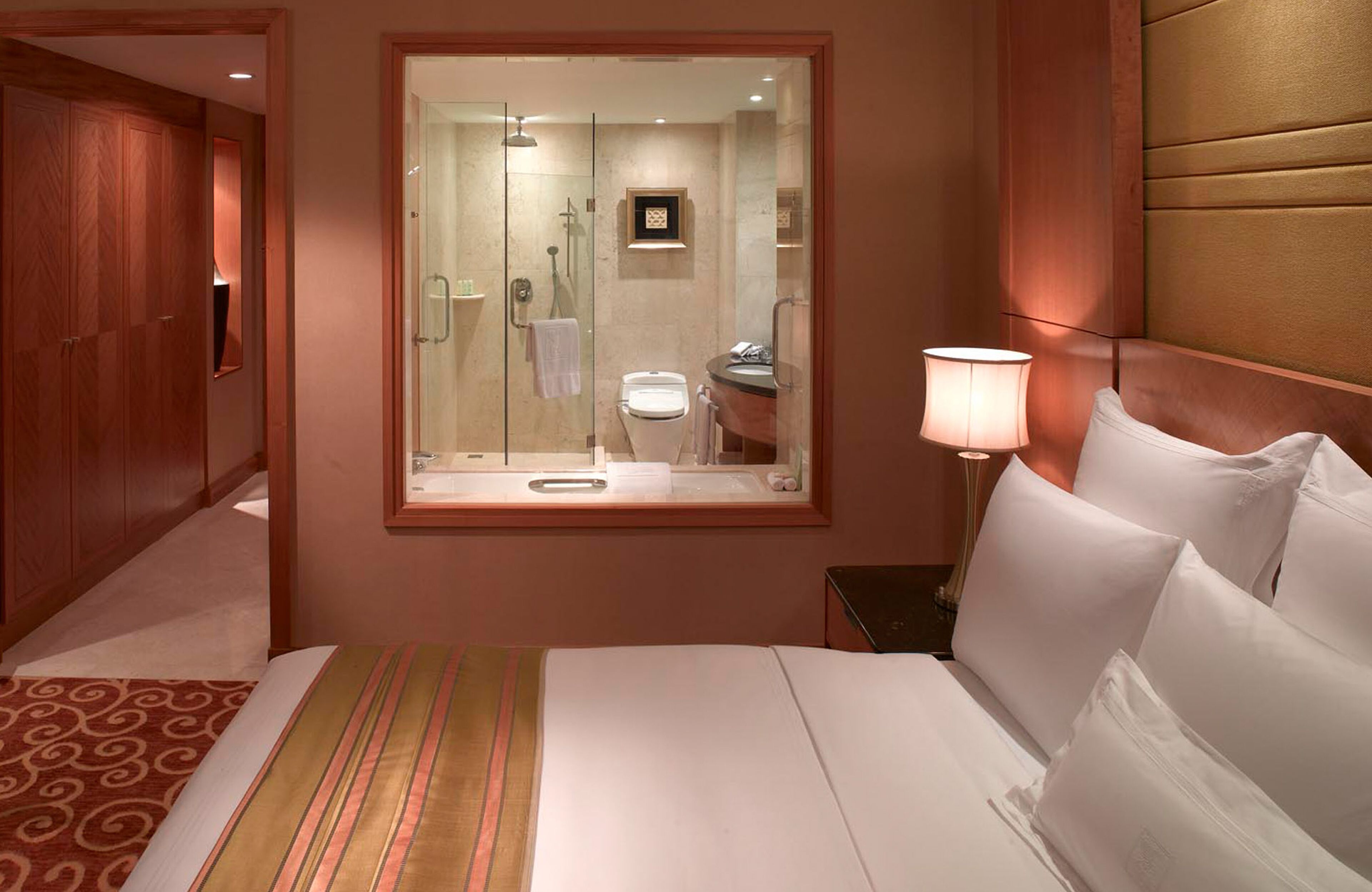 A hotel room with a large bed, a bedside lamp, a mirror, a bathroom visible through the mirror, and a wooden wardrobe.