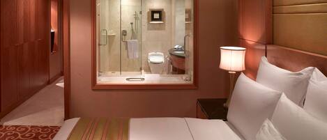 A hotel room with a large bed, a bedside lamp, a mirror, a bathroom visible through the mirror, and a wooden wardrobe.