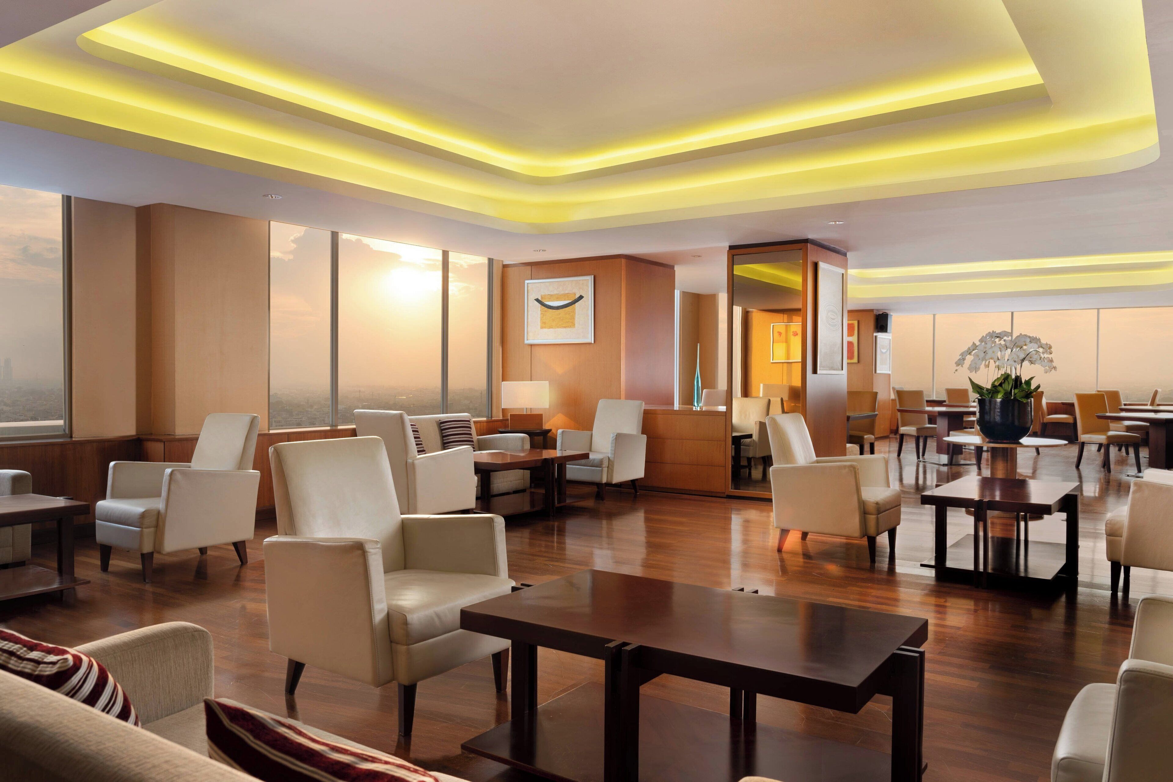 A spacious hotel lobby with multiple seating areas, including sofas and chairs, arranged around a central coffee table. There is a reception desk with a large potted plant and a framed artwork on the wall.
