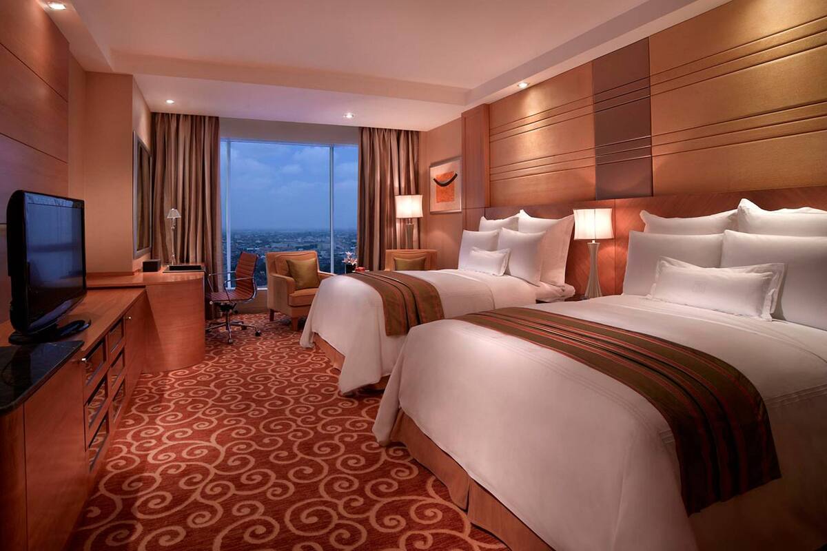 A hotel room with two beds, a TV, a desk, and a view of the city.