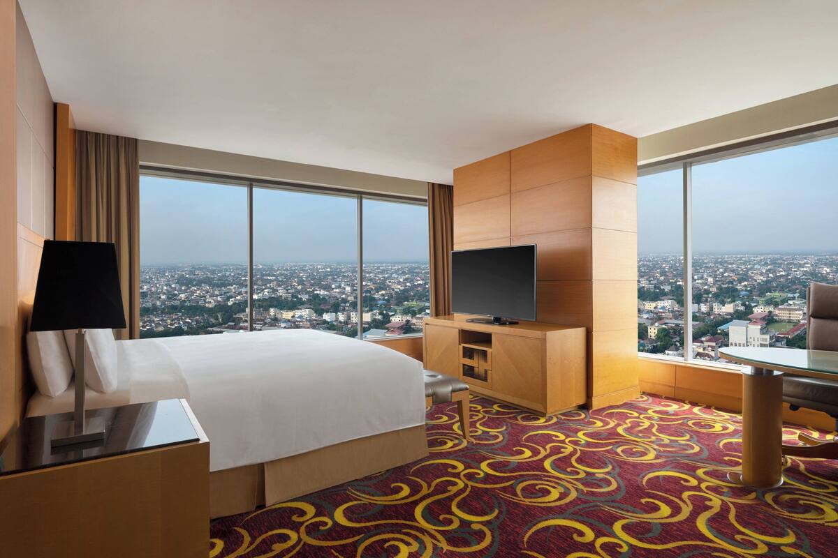 A hotel room with a large bed, a TV, a desk, and a view of a cityscape through large windows.