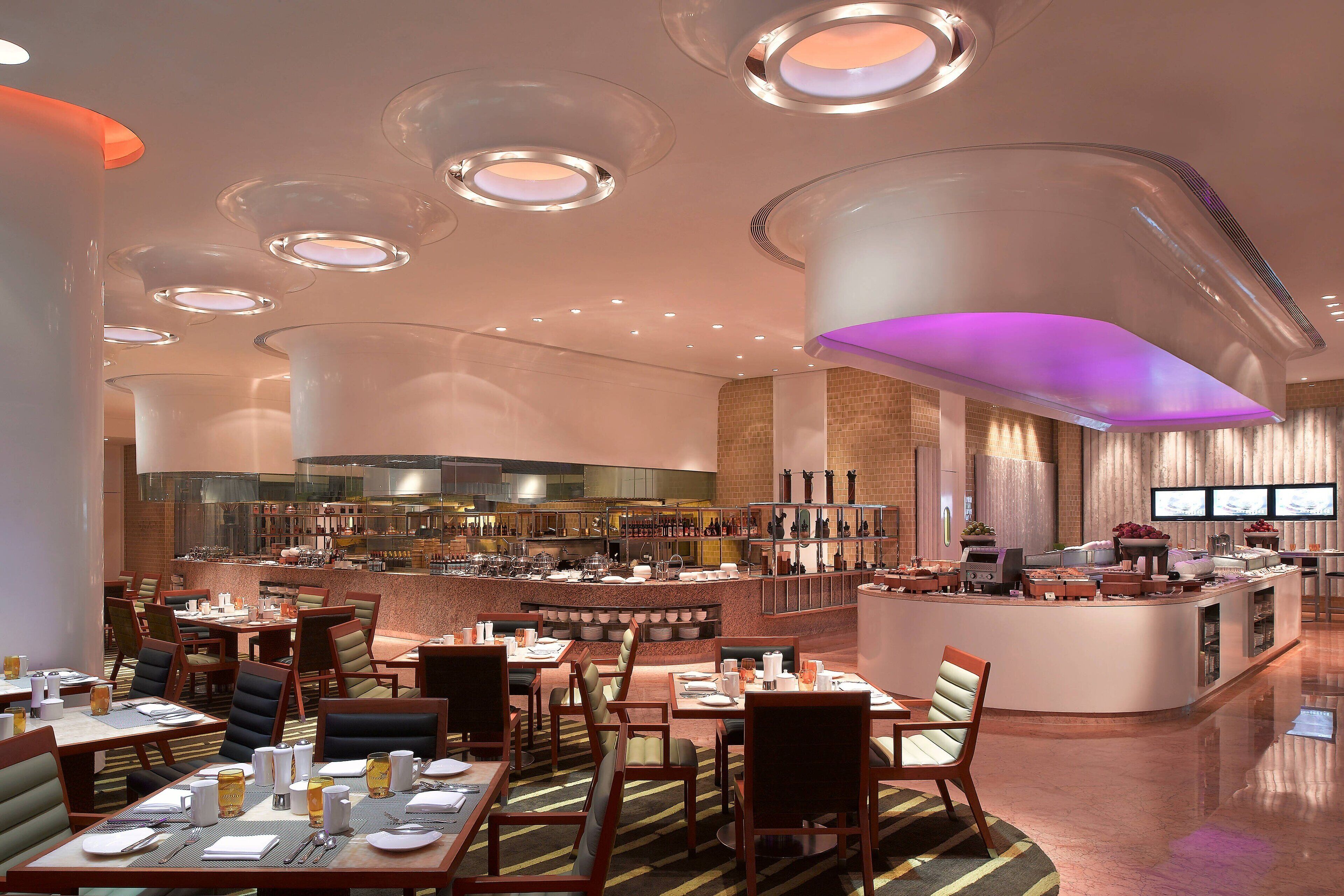 A modern restaurant with round tables, a central bar, and a seating area with cushioned chairs.