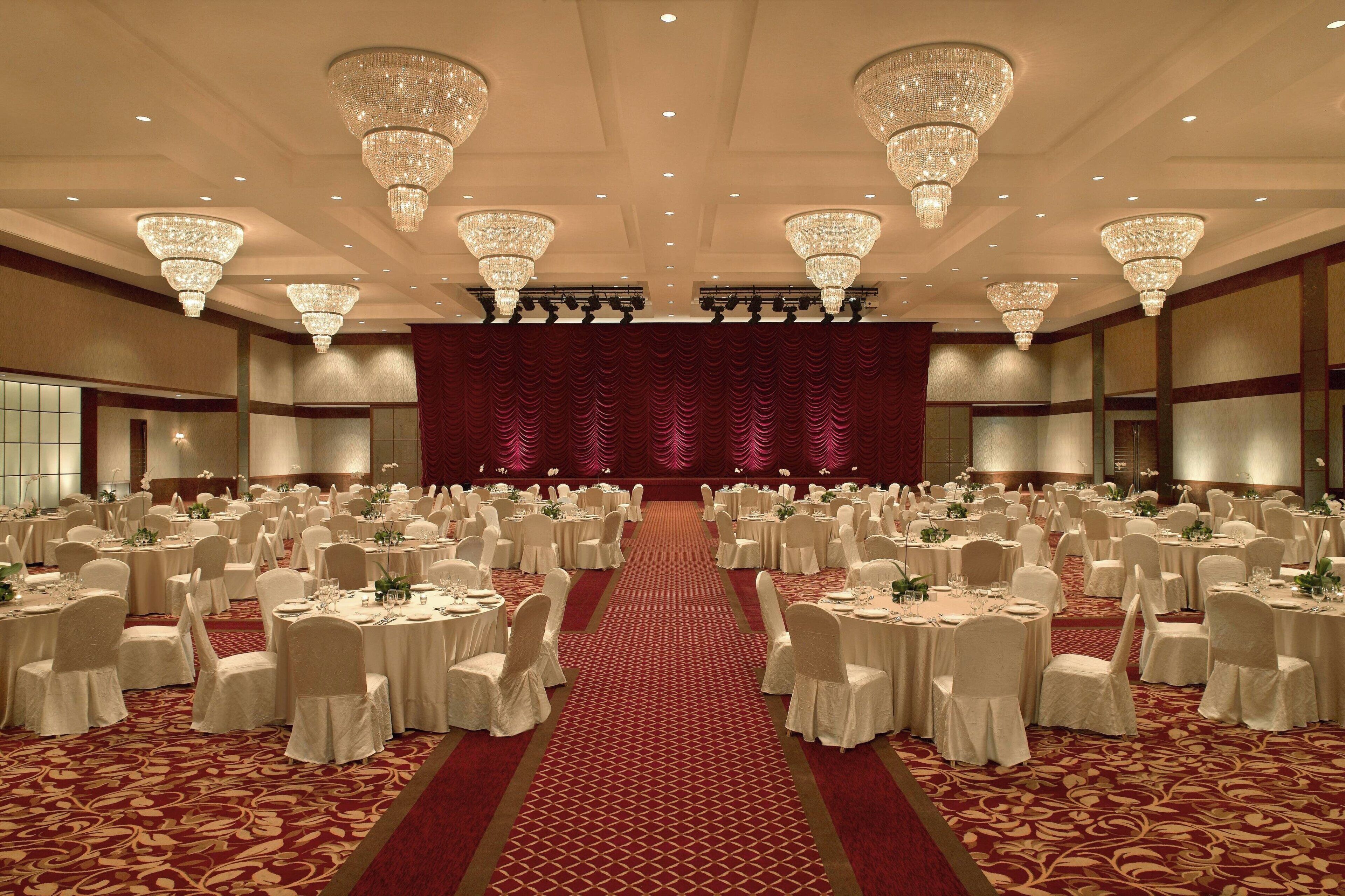 A banquet hall with round tables, white tablecloths, and chairs, a red carpet, and chandeliers.