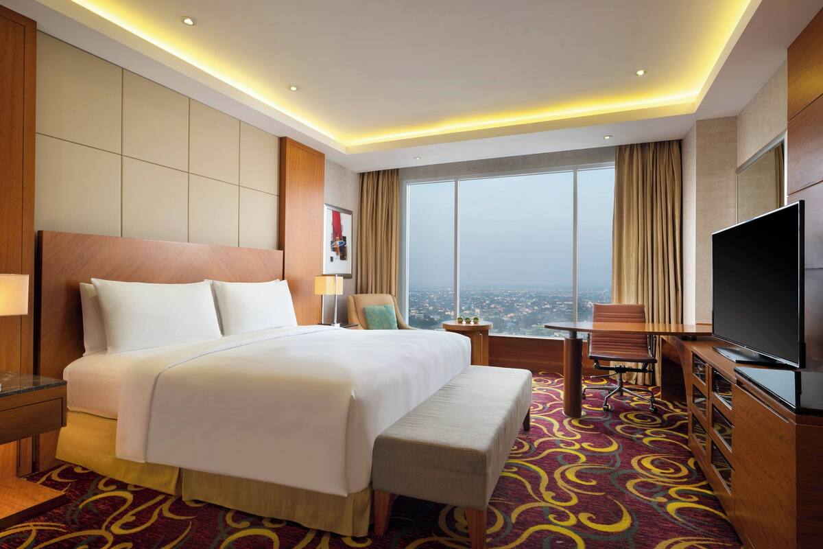 A hotel room with a large bed, a TV, a desk, and a view of the city.