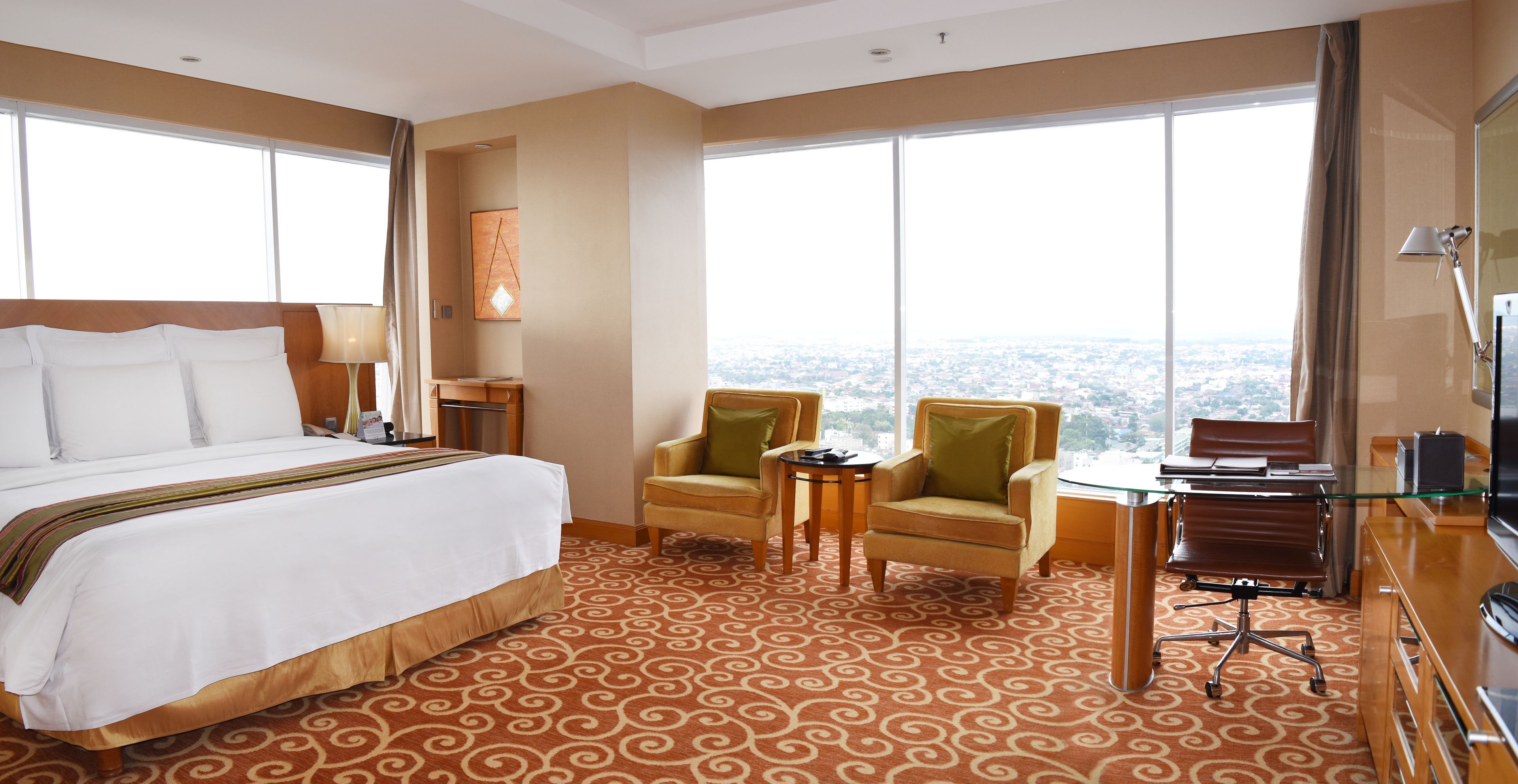 Habitación de hotel con una cama grande, dos sillones, una mesita y un escritorio con una silla. La habitación tiene ventanas amplias que ofrecen vistas a la ciudad.
