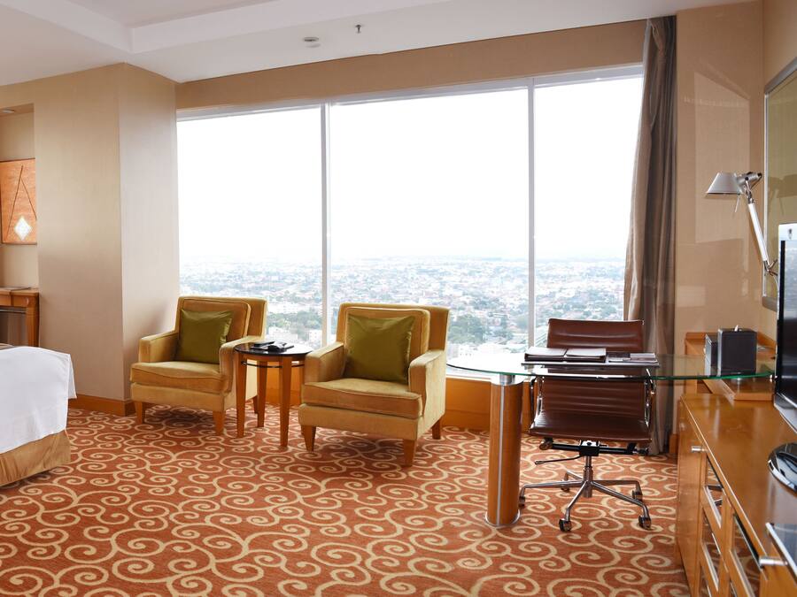 Habitación de hotel con una cama grande, dos sillones, una mesita y un escritorio con una silla. La habitación tiene ventanas amplias que ofrecen vistas a la ciudad.