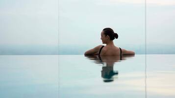 A person in a black swimsuit sitting in a clear glass infinity pool with a view of the horizon.