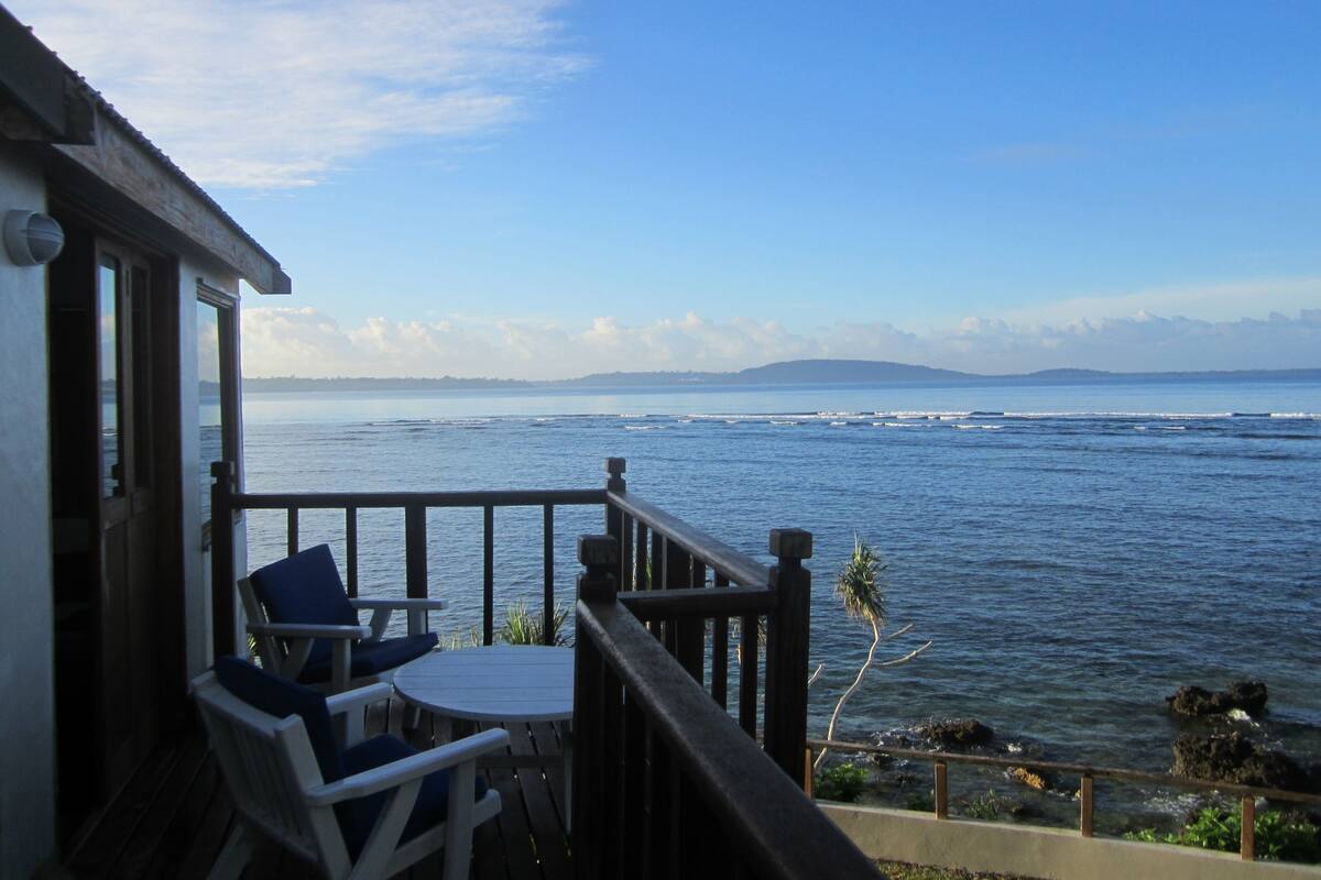 Une terrasse en bois avec une table et des chaises, donnant sur une mer calme aux vagues douces, un ciel dégagé et une terre lointaine.