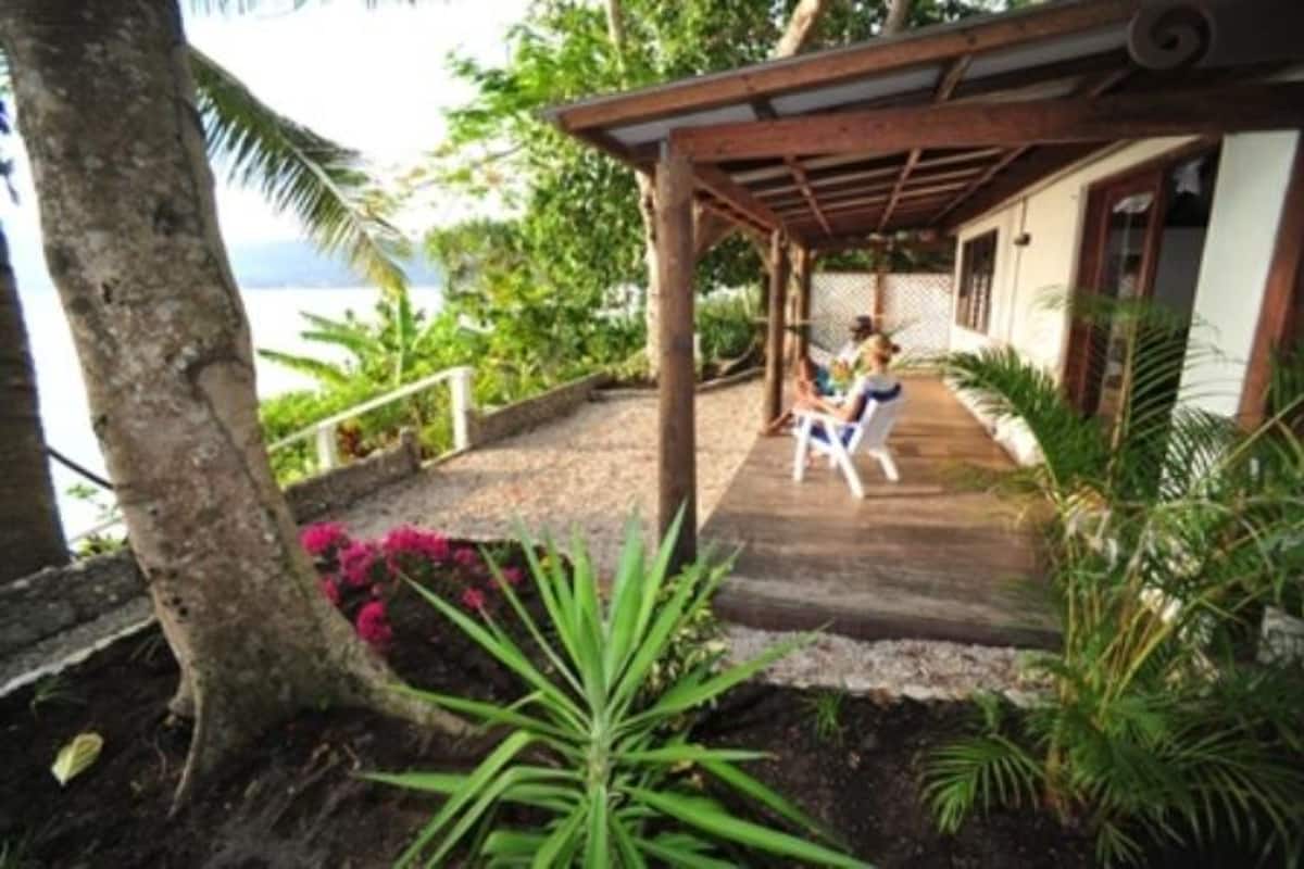 Une véranda en bois, ouverte sur l’océan, entourée de plantes tropicales et d’un arbre.