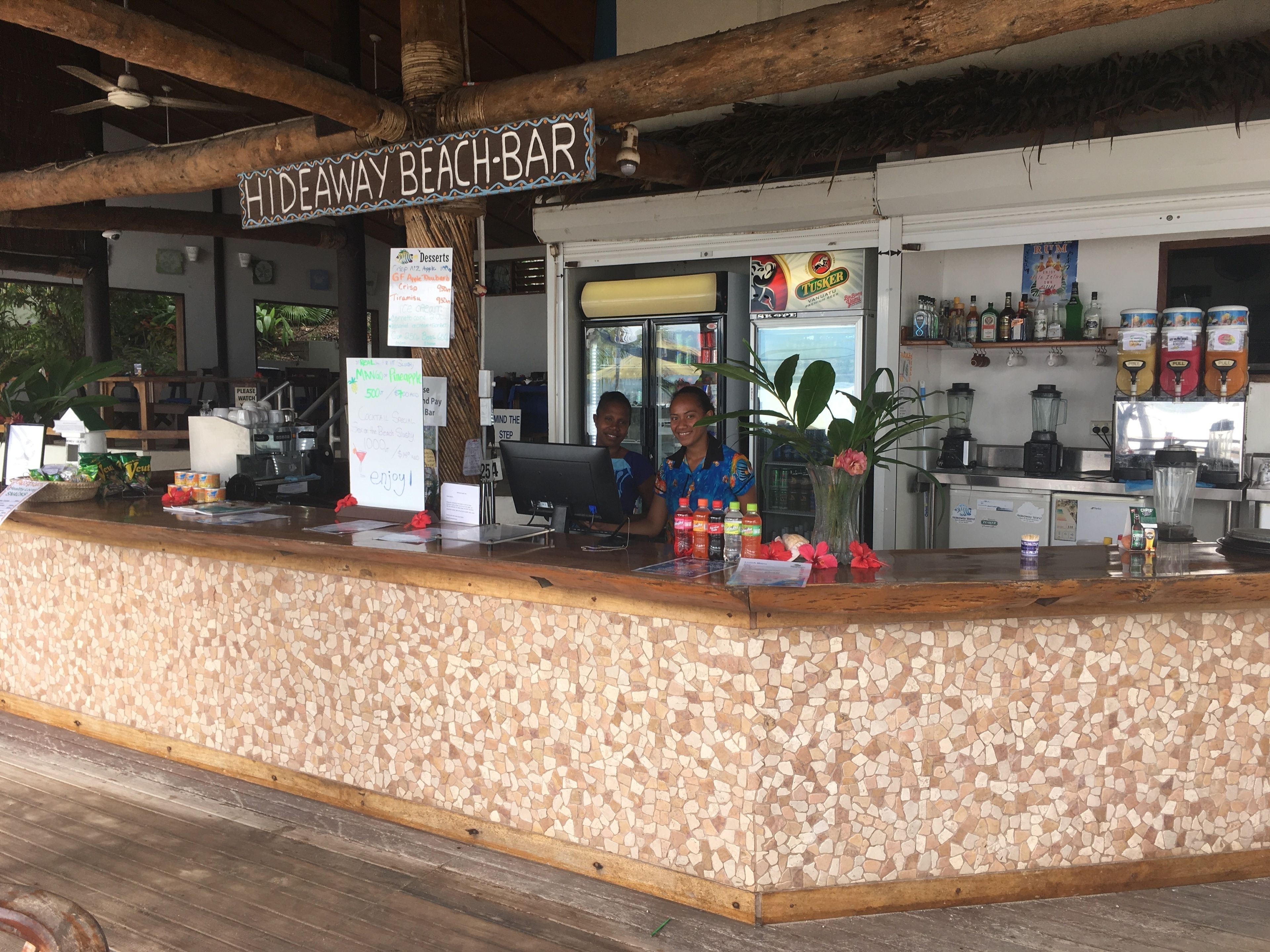 Un bar sulla spiaggia con un bancone di legno, una bacheca dei menu e un cartello sopra il bar.