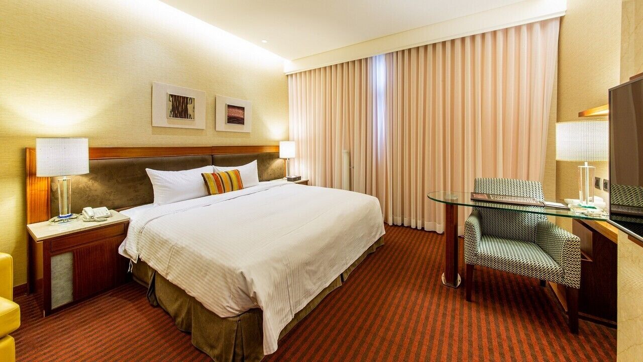 A hotel room with a large bed, a desk with a chair, a lamp, and a television.