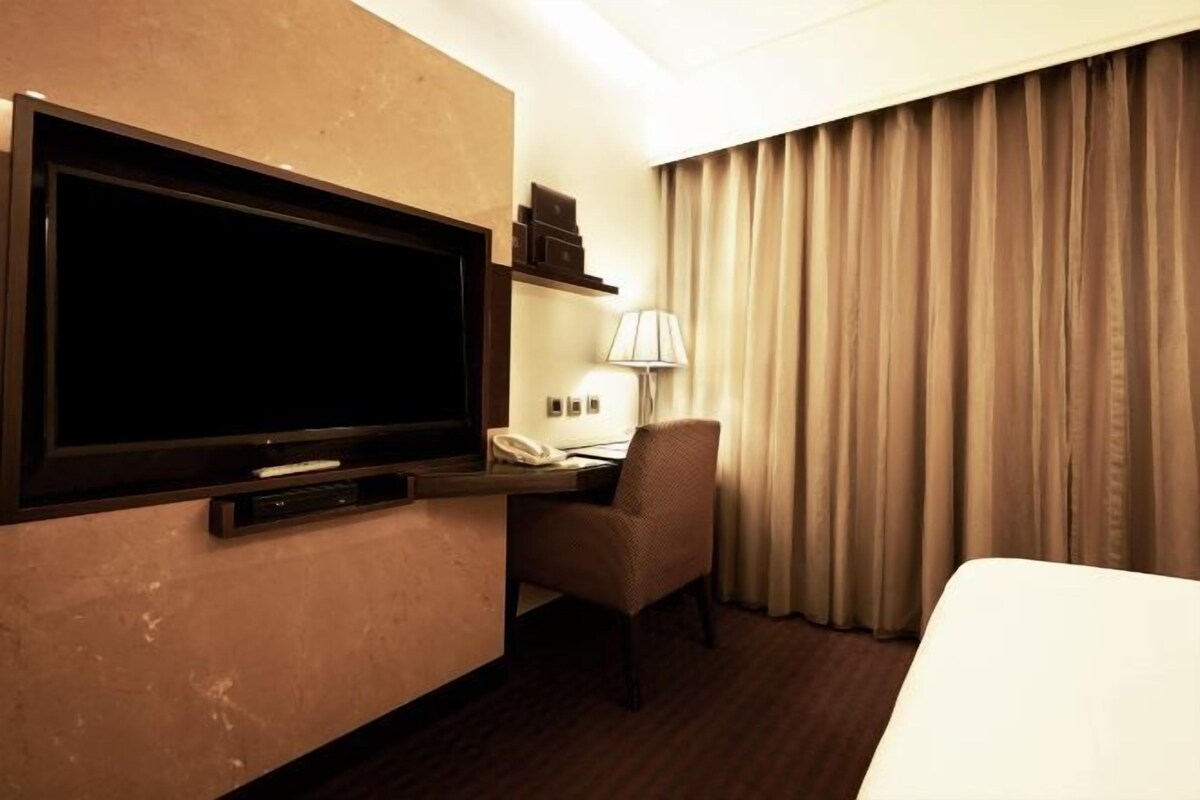 Ett hotellrum med en stor platt-TV som är monterad på väggen, ett skrivbord med en lampa, en stol och en säng.