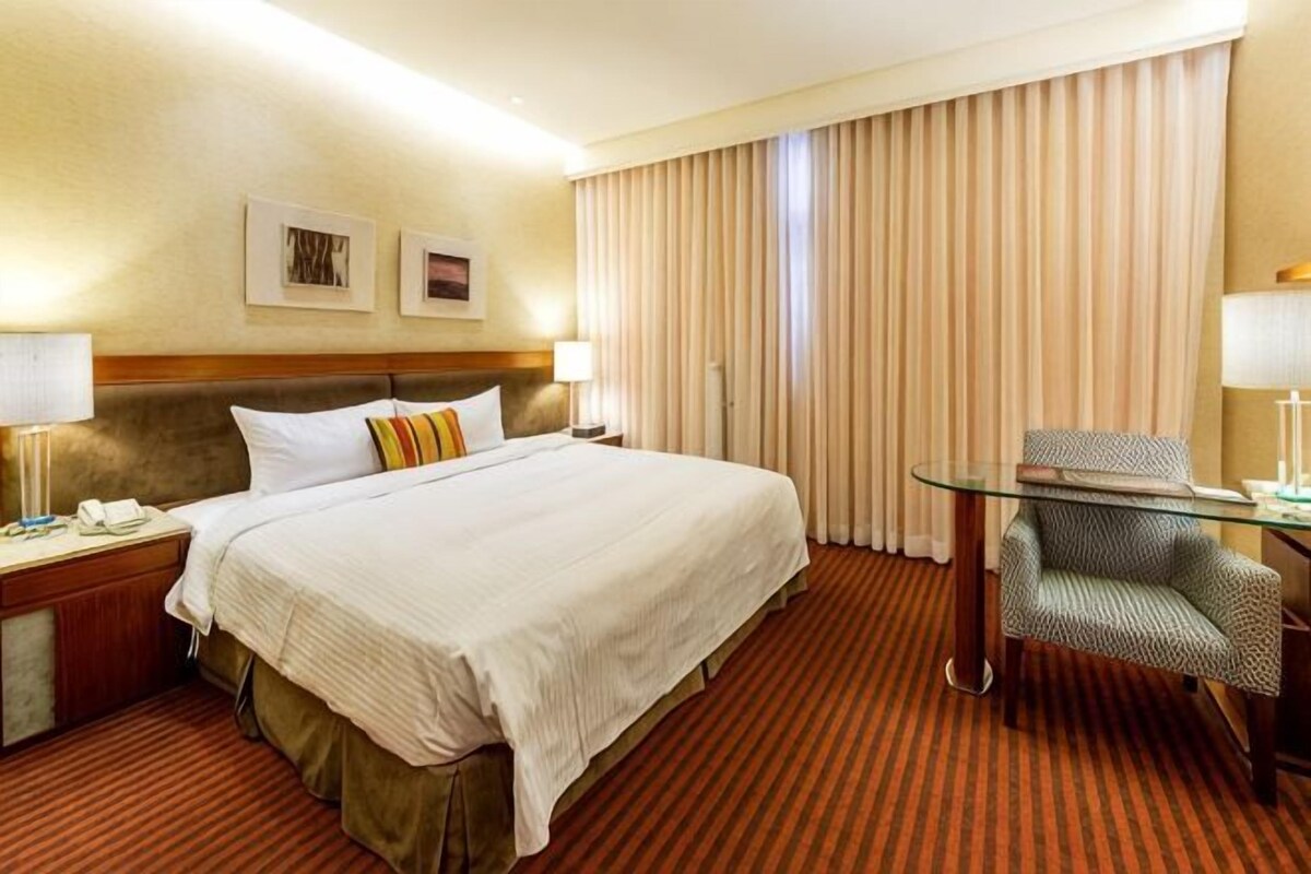 Une chambre d’hôtel avec un grand lit, un bureau avec une chaise, une table de chevet et une fenêtre avec des rideaux.