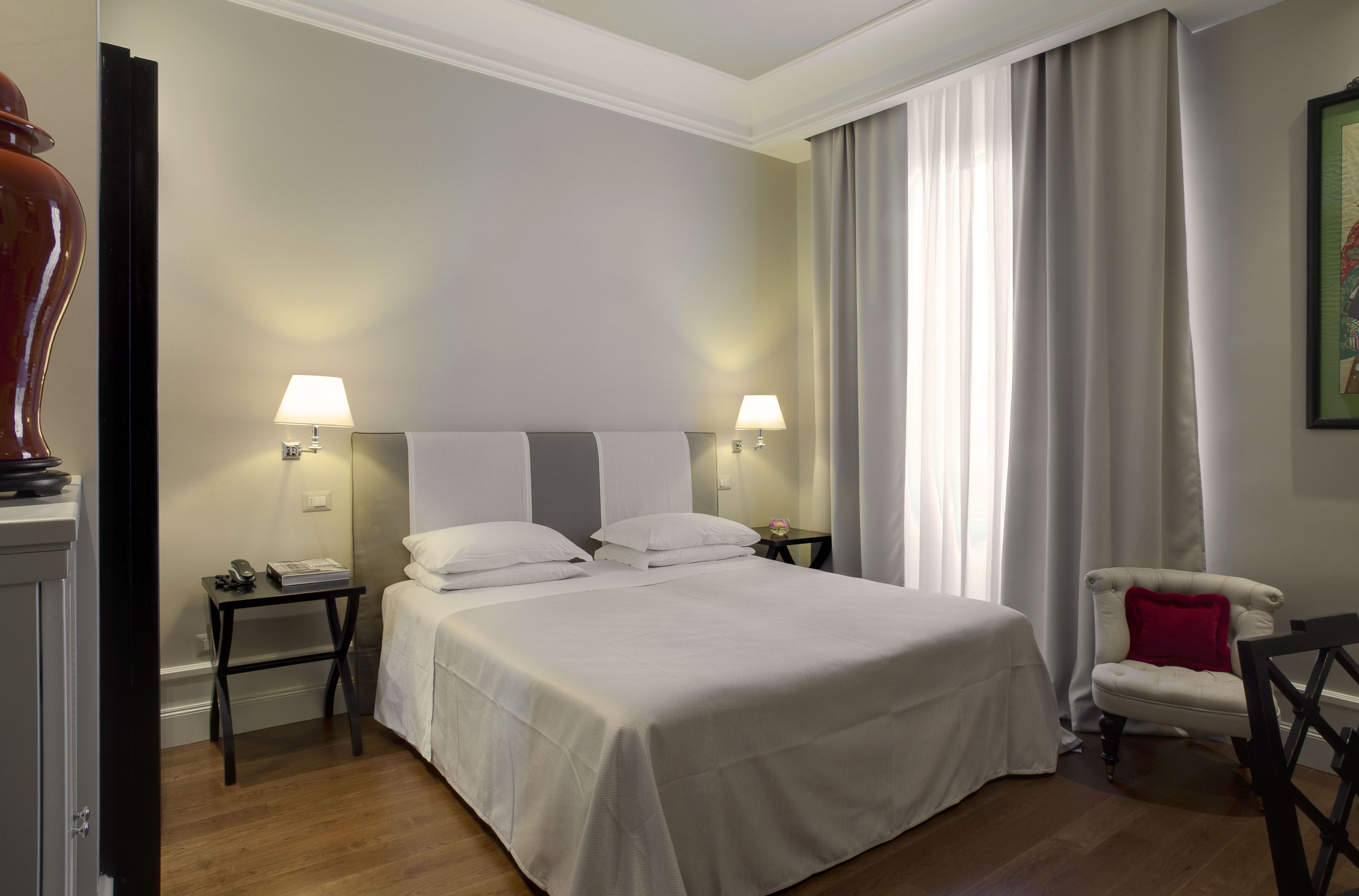 A hotel room with a large bed, two bedside tables with lamps, a chair with a red cushion, and a painting on the wall.