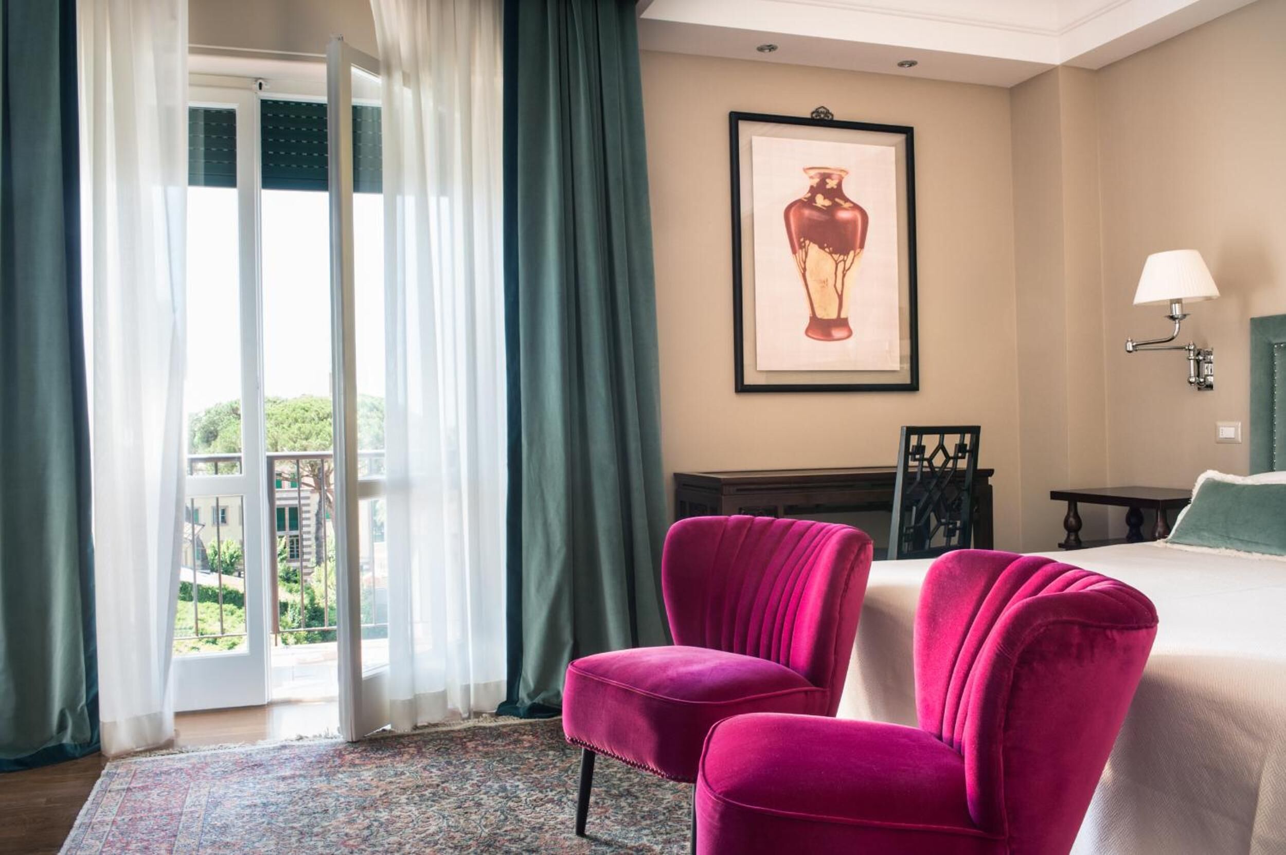 Habitación de hotel con una cama, dos sillas rosas, una obra de arte enmarcada en la pared, una mesita de noche y vistas a través de una puerta de vidrio.
