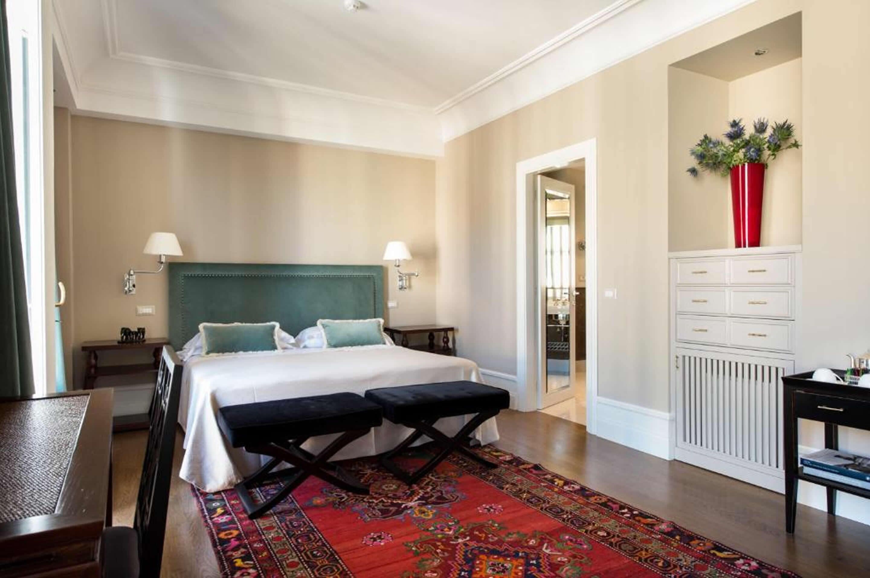 A hotel room with a bed, two bedside tables, a nightstand, a red vase, and a patterned rug.