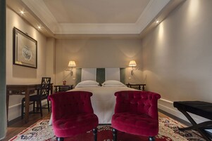 Una habitación de hotel con una cama grande, dos sillones de terciopelo rojo, un escritorio de madera y una alfombra estampada.