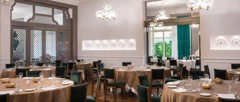 A formal dining room with round tables, green upholstered chairs, and chandeliers.