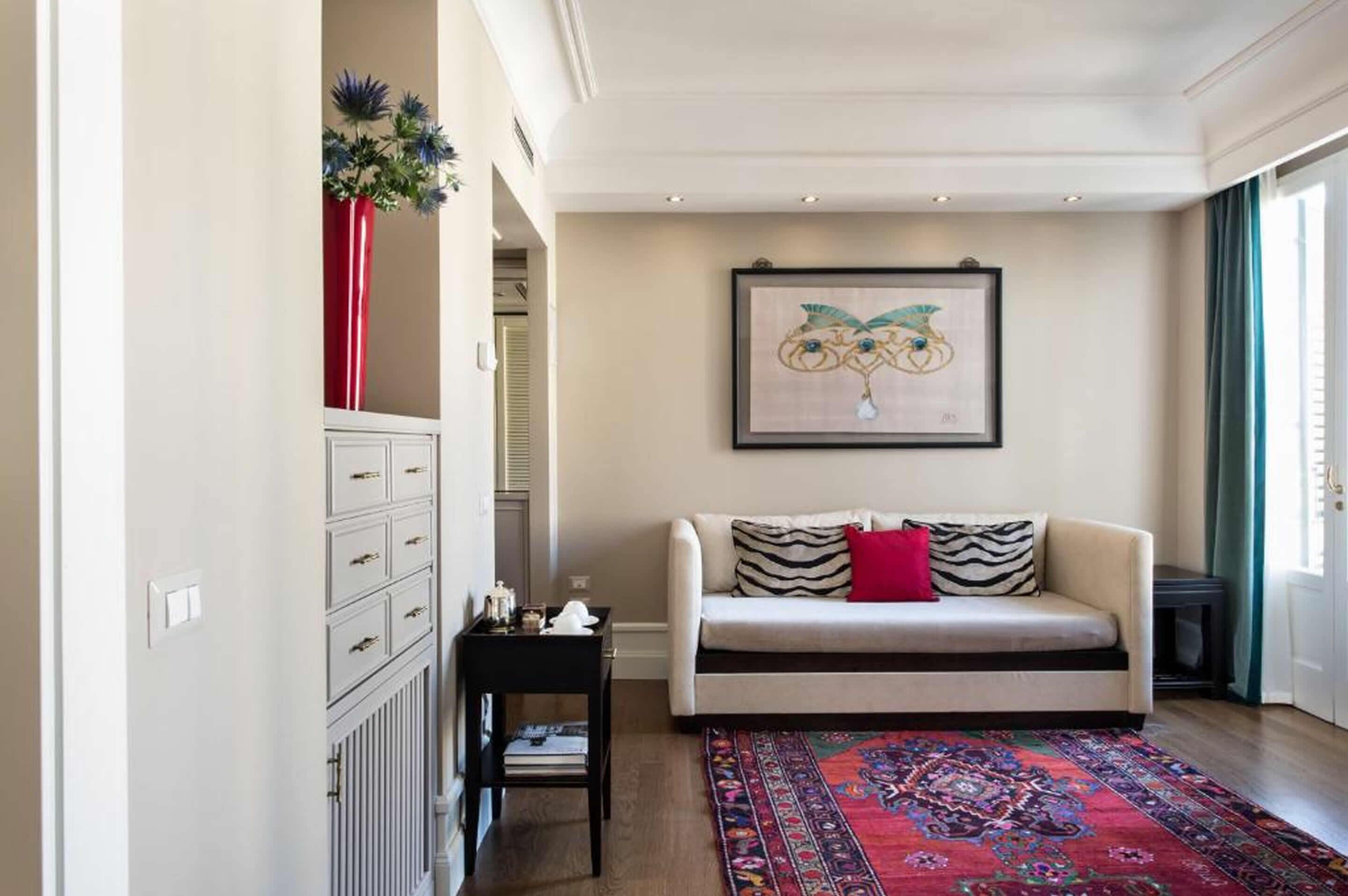 Un salon avec un canapé beige, un tapis à motifs, un vase rouge contenant des fleurs et une œuvre d’art encadrée accrochée au mur.