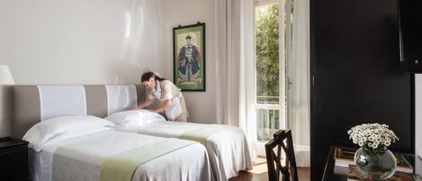 Habitación de hotel con dos camas, un ventanal con cortinas, un cuadro en la pared y una mesa con un jarrón de flores.