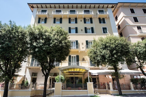 A multi-story hotel with a central entrance, balconies, and trees in the foreground.
