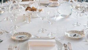 A formal dining table set with multiple wine glasses, plates, and cutlery, with a decorative centerpiece and candles in the background.