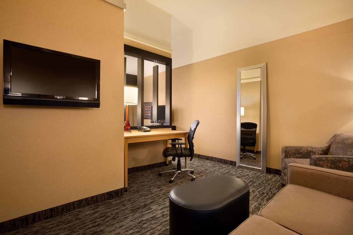 Habitación de hotel con televisión de pantalla plana en la pared, un escritorio con una silla, un sofá y un espejo grande.