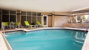 A swimming pool with lounge chairs and a small table, surrounded by large windows and a stone wall.