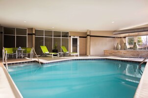 A swimming pool with lounge chairs and a small table, surrounded by large windows and a stone wall.