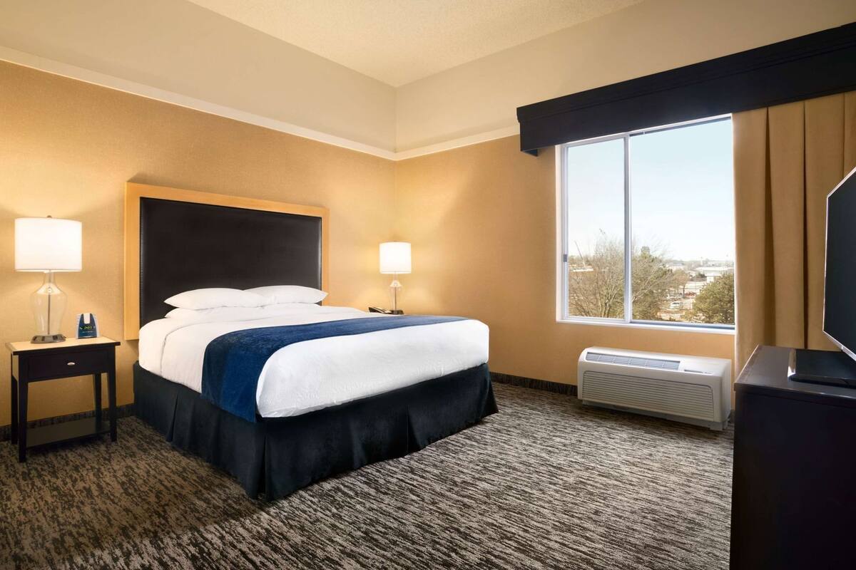 Habitación de hotel con cama, dos lámparas de noche, ventana con vistas, equipo de aire acondicionado y televisor sobre un mueble.
