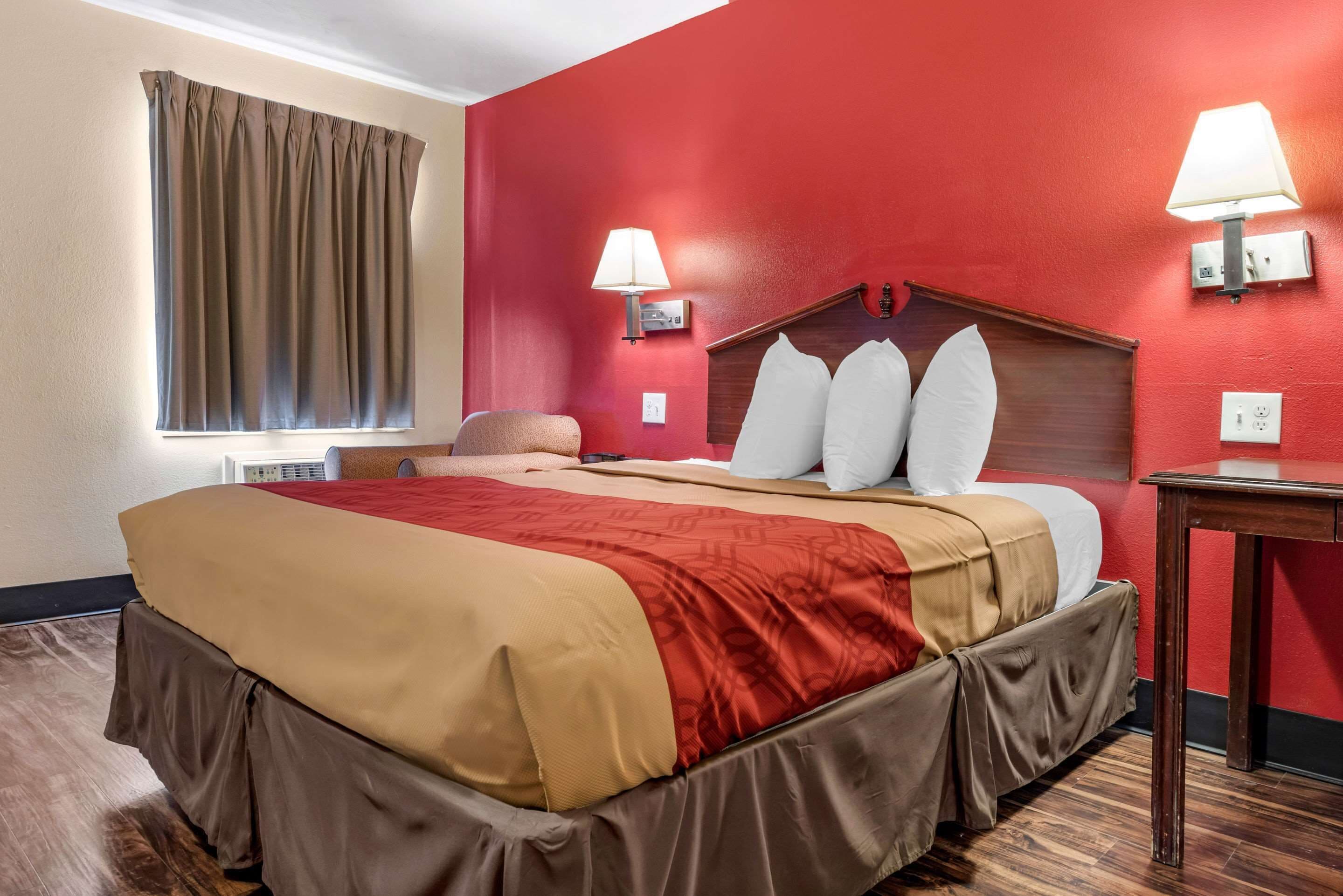 Habitación de hotel con cama, paredes rojas, dos lámparas de pared, una ventana con cortinas y piso de madera.