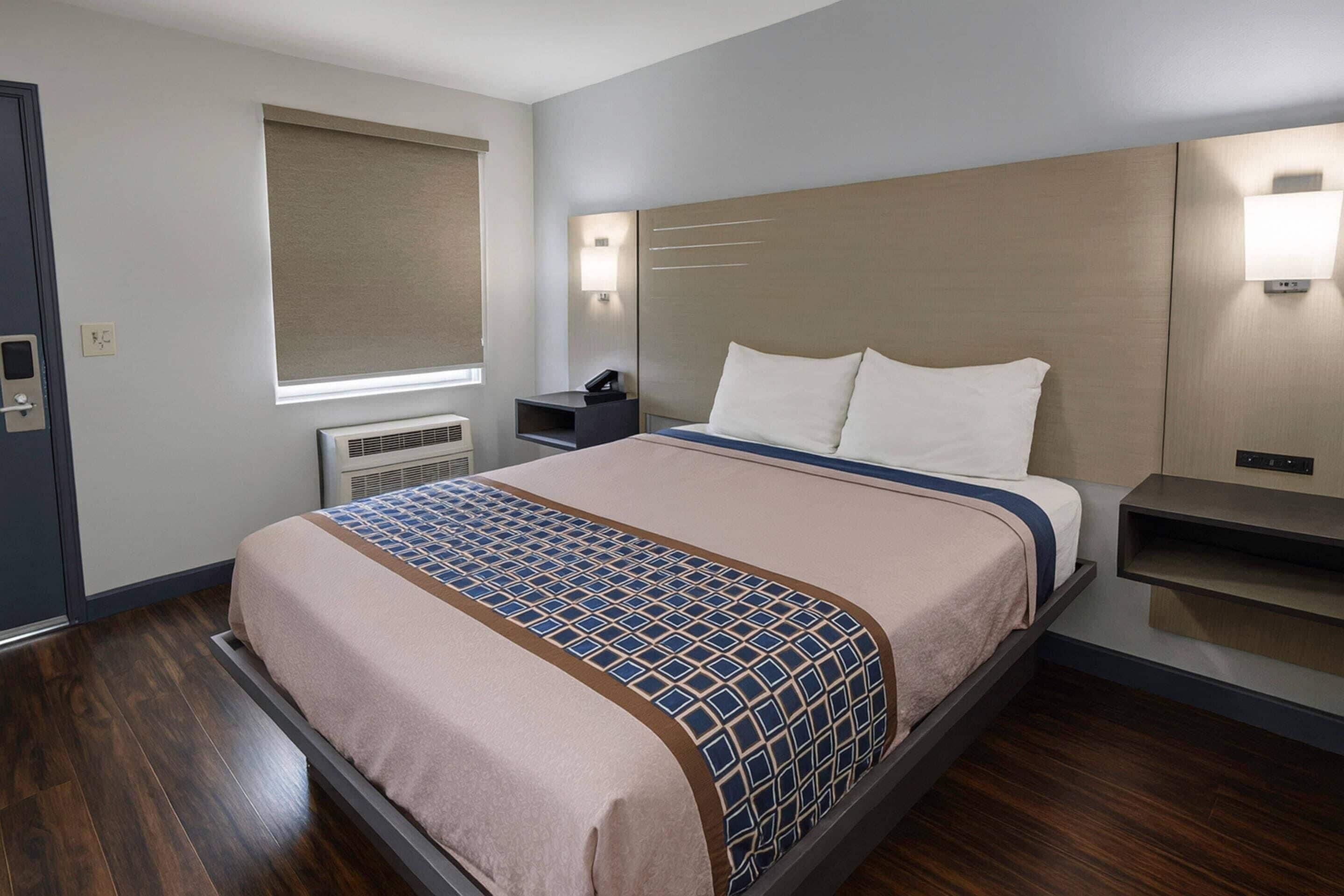 A hotel room with a bed, bedside tables, a window with blinds, a wall-mounted light, and a thermostat.