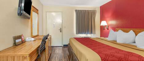 Quarto de hotel com cama, escrivaninha, cadeira, televisão, janela com cortinas e luminária.