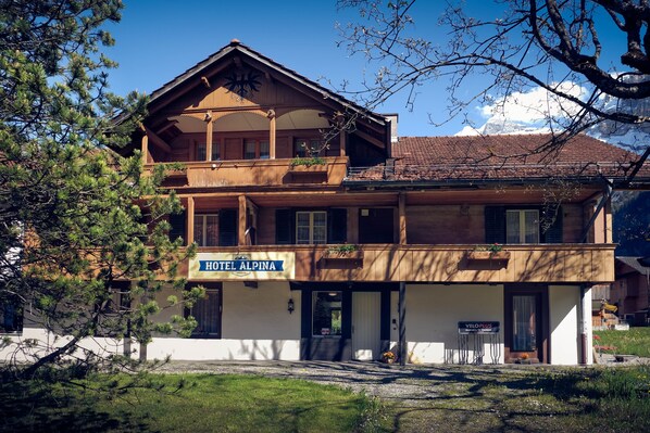 Un bâtiment en bois à deux étages, doté d’un balcon, sur lequel on peut lire l’inscription « HÔTEL ALPINA », et une enseigne indiquant « VELO PLUS », ce qui signale la présence d’un service à proximité.