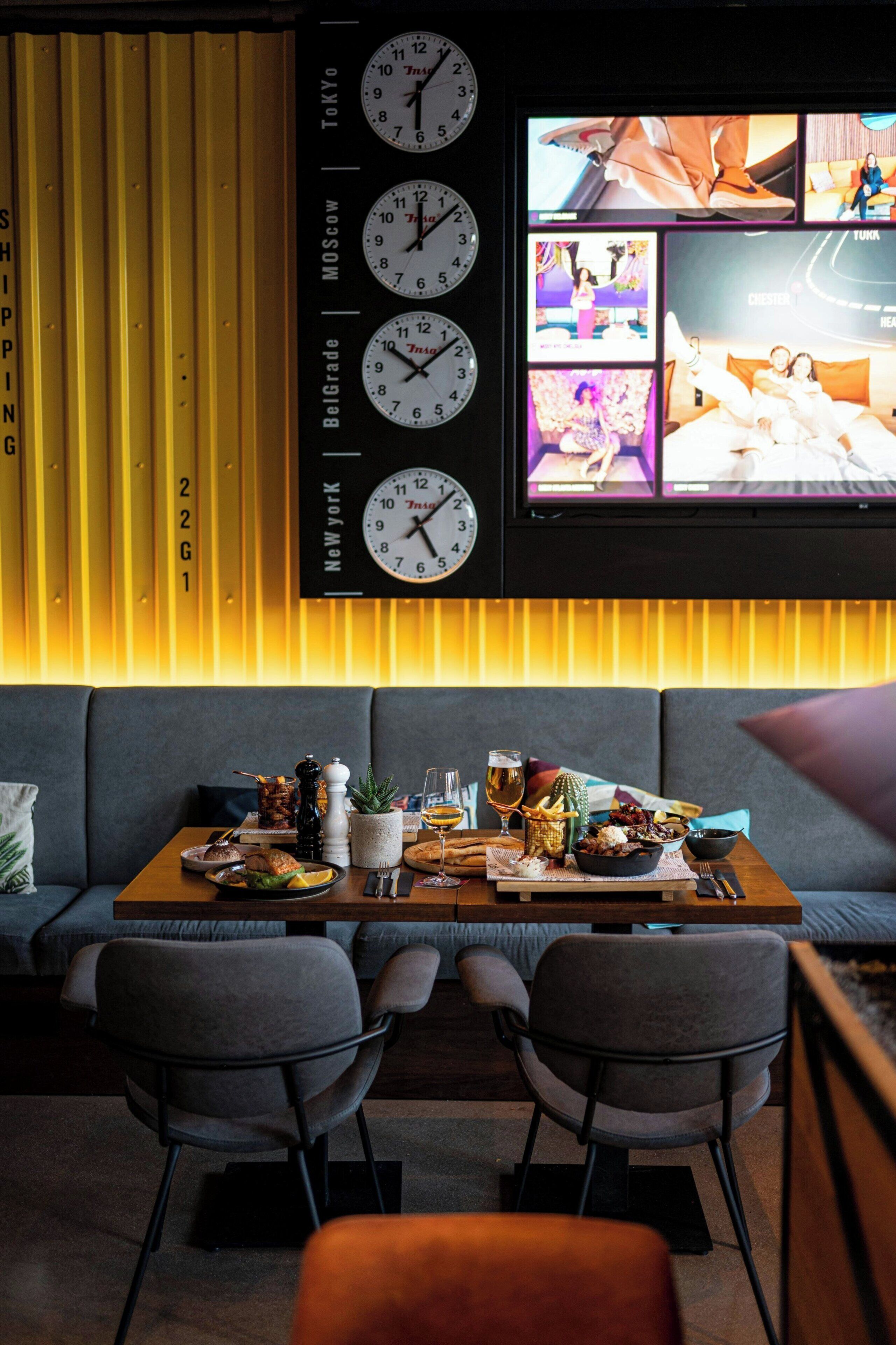 A modern interior with a sofa, two chairs, a wooden table with food, and a wall clock displaying multiple time zones.