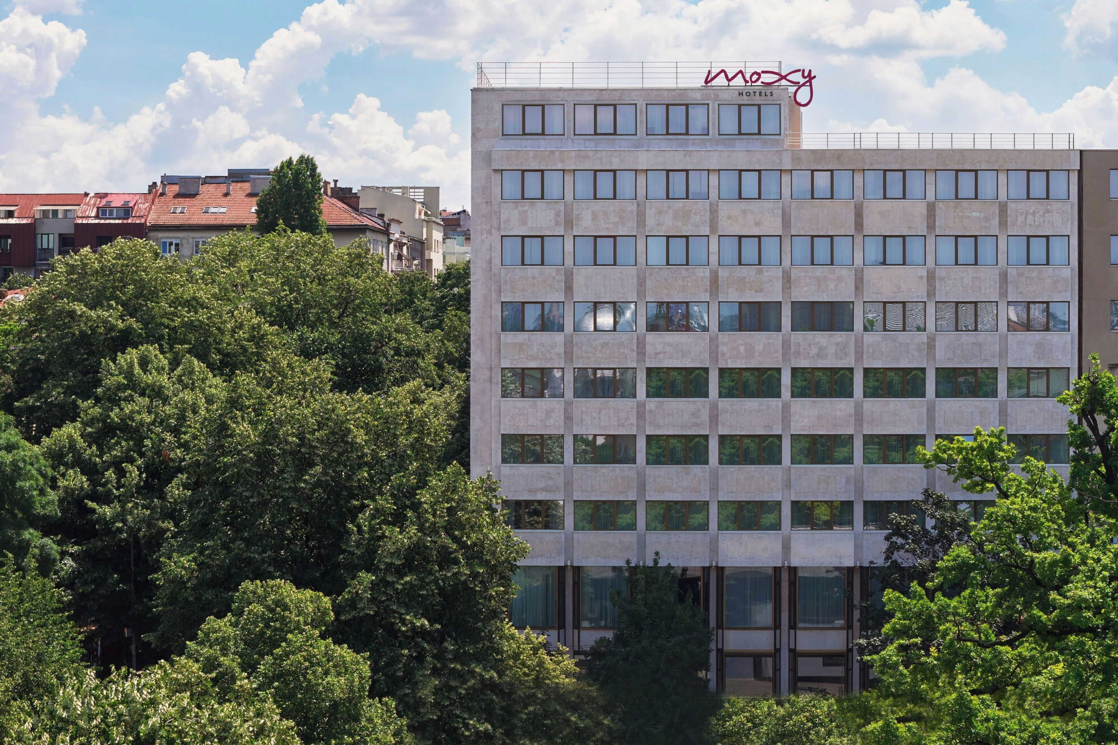 A modern hotel building with multiple floors and a flat roof, surrounded by lush green trees.