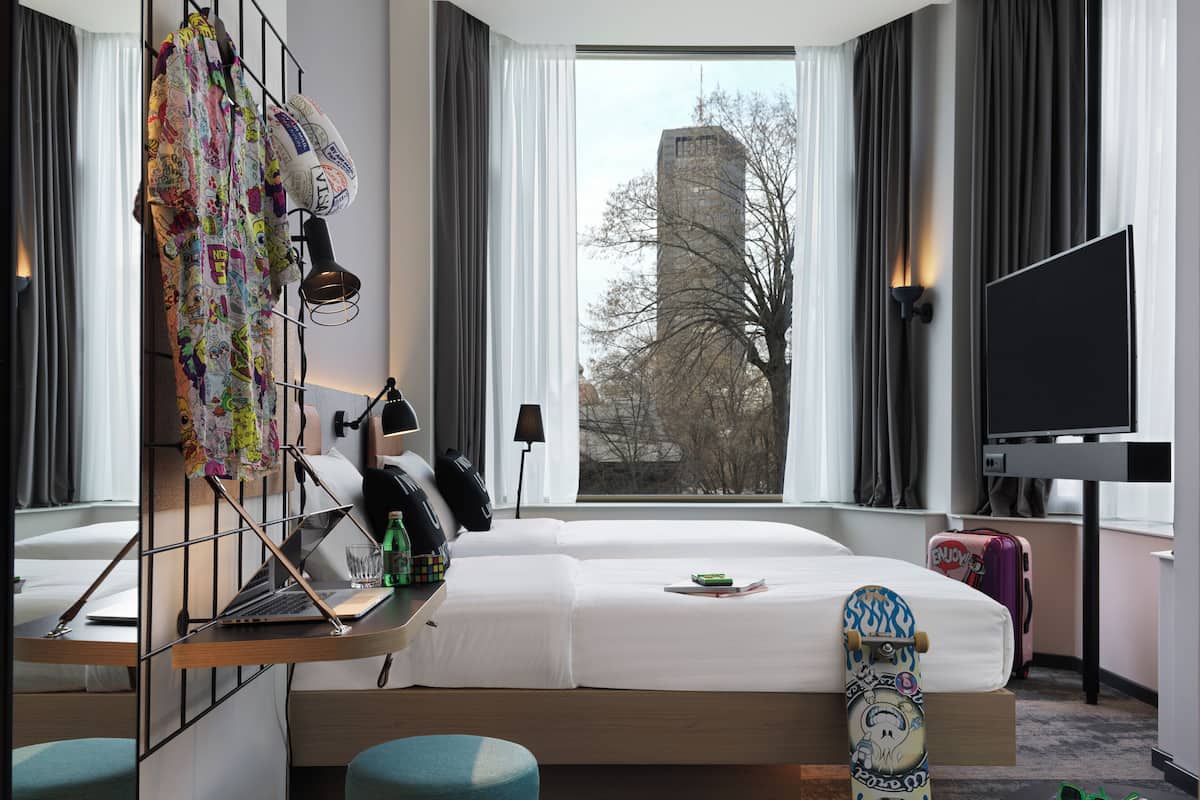A modern hotel room with a large bed, a desk with a laptop, a TV, and a view of a building through the window.