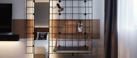 A hotel room with a bed, a bedside table, a mirror, a wall-mounted shelf, and a small stool.